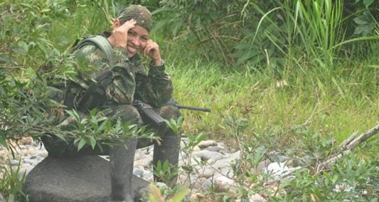 Olga, smiling and sitting on a rock, wearing camouflage and carrying a gun. 